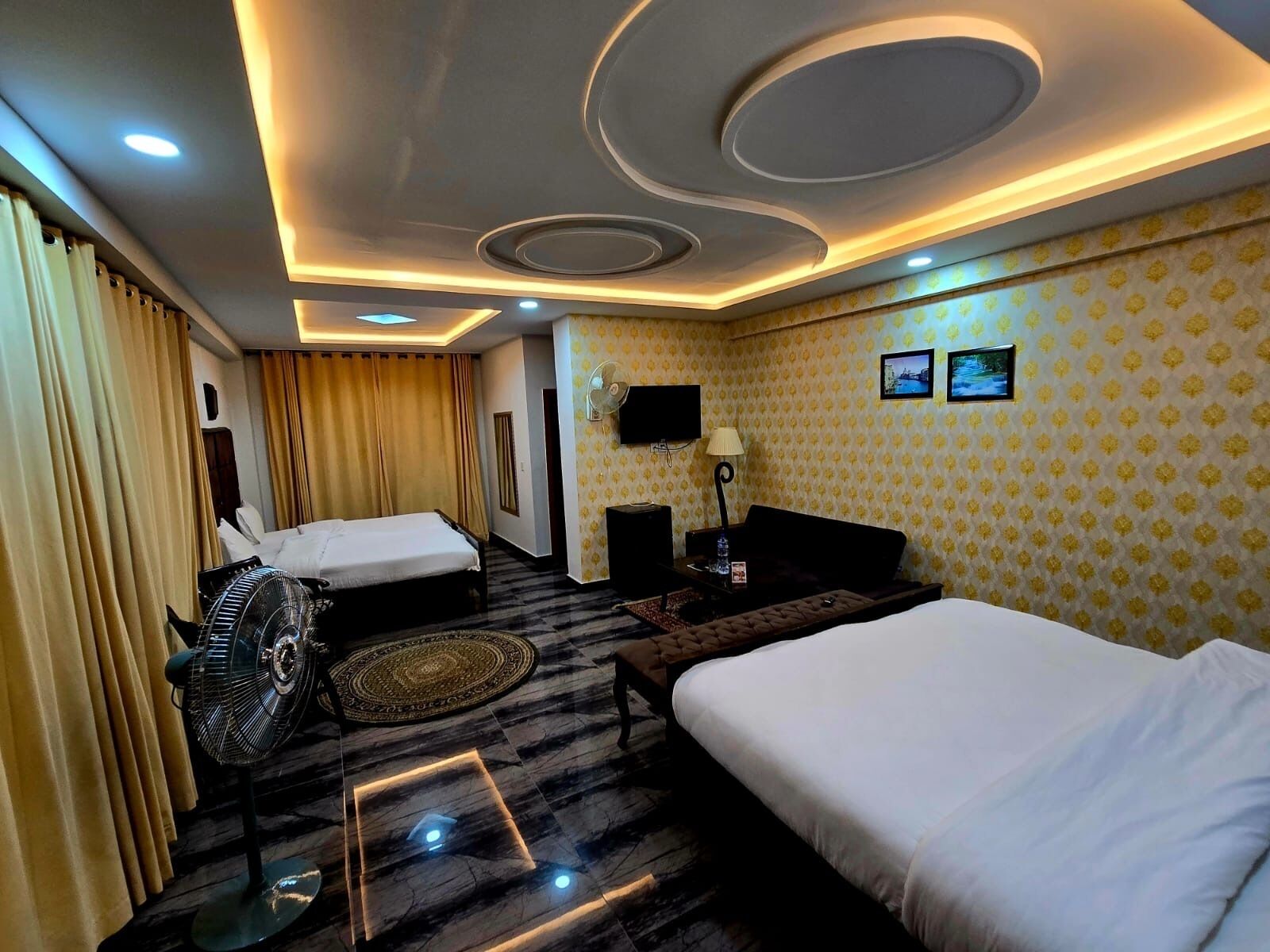 A hotel room with two beds, a flat-screen TV, a desk with a lamp, and a patterned wallpaper.