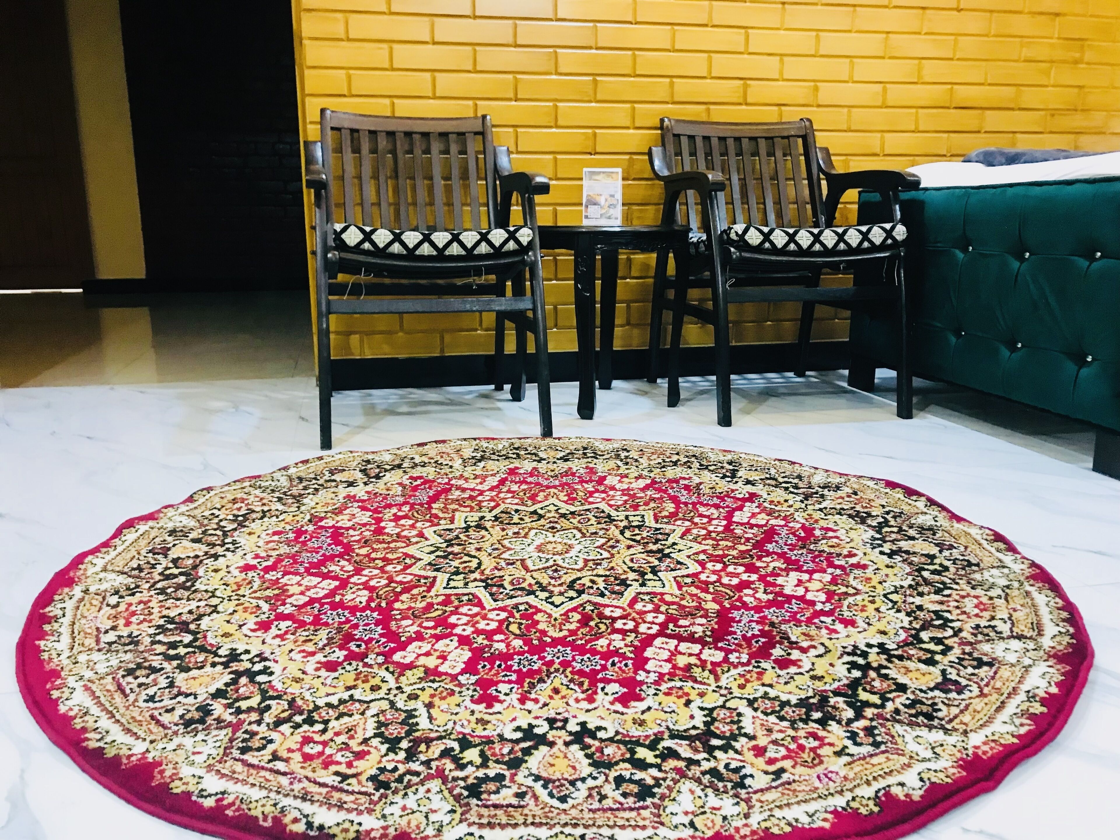 Ein Zimmer mit einem runden Teppich mit Muster, zwei Holzstühle mit Kissen, einem kleinen Tisch und einem grünen, gepolsterten Sofa an einer gelben Backsteinwand.
