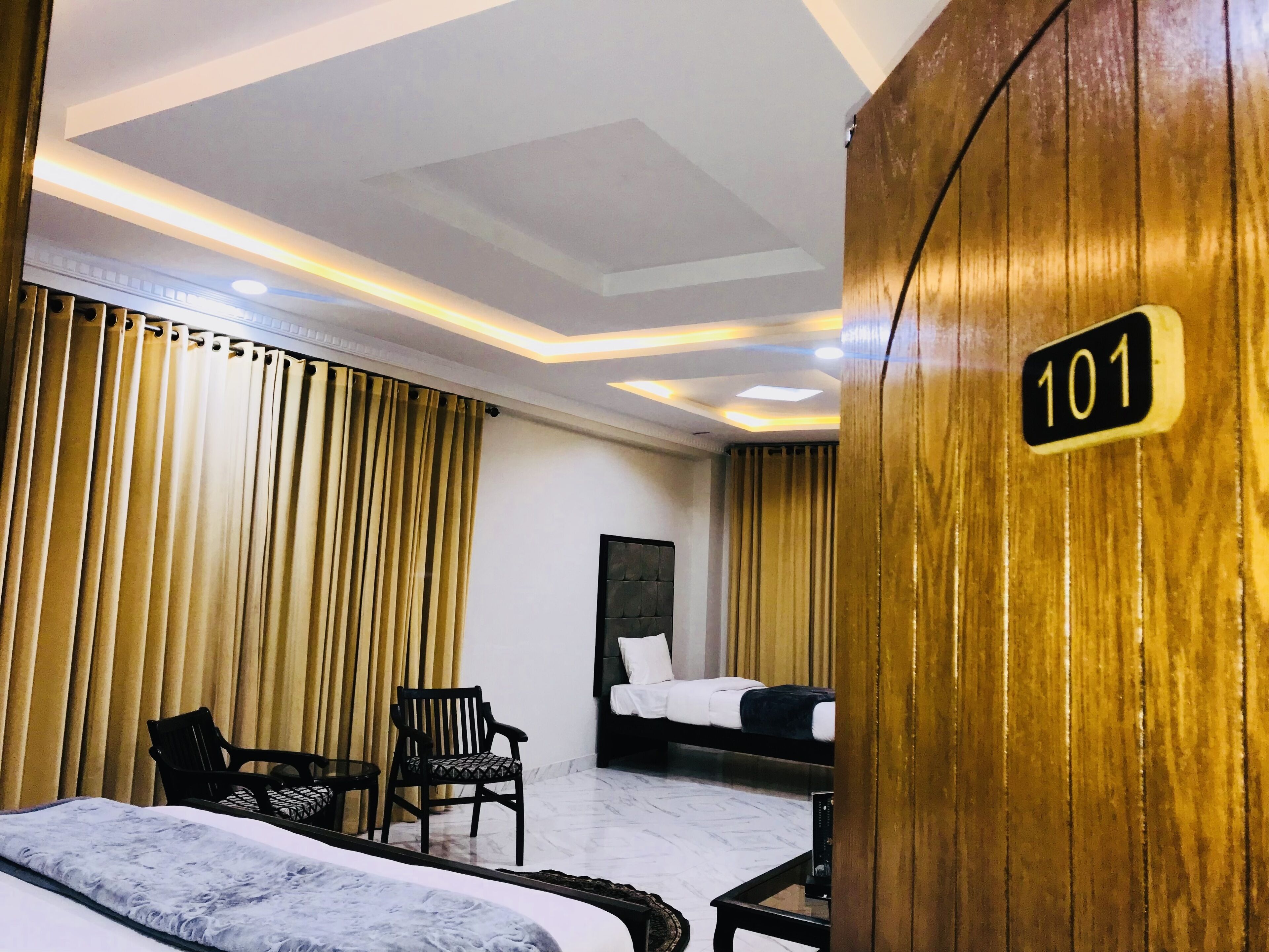 A hotel room with two beds, a wooden accent wall, a number sign '101', and a modern ceiling design.