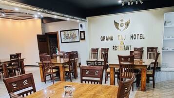 A spacious dining area with a long wooden table surrounded by chairs, a high ceiling with modern lighting, and a sign for 'Grand Taj Hotel & Restaurant'.