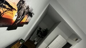 Une chambre à coucher avec un lit, une table de chevet ornée d’une plante, un tableau accroché au mur et une porte donnant sur une autre pièce.