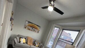 Un salon avec un canapé gris, une table basse noire, un tapis jaune et gris, un ventilateur de plafond et une fenêtre avec des stores.