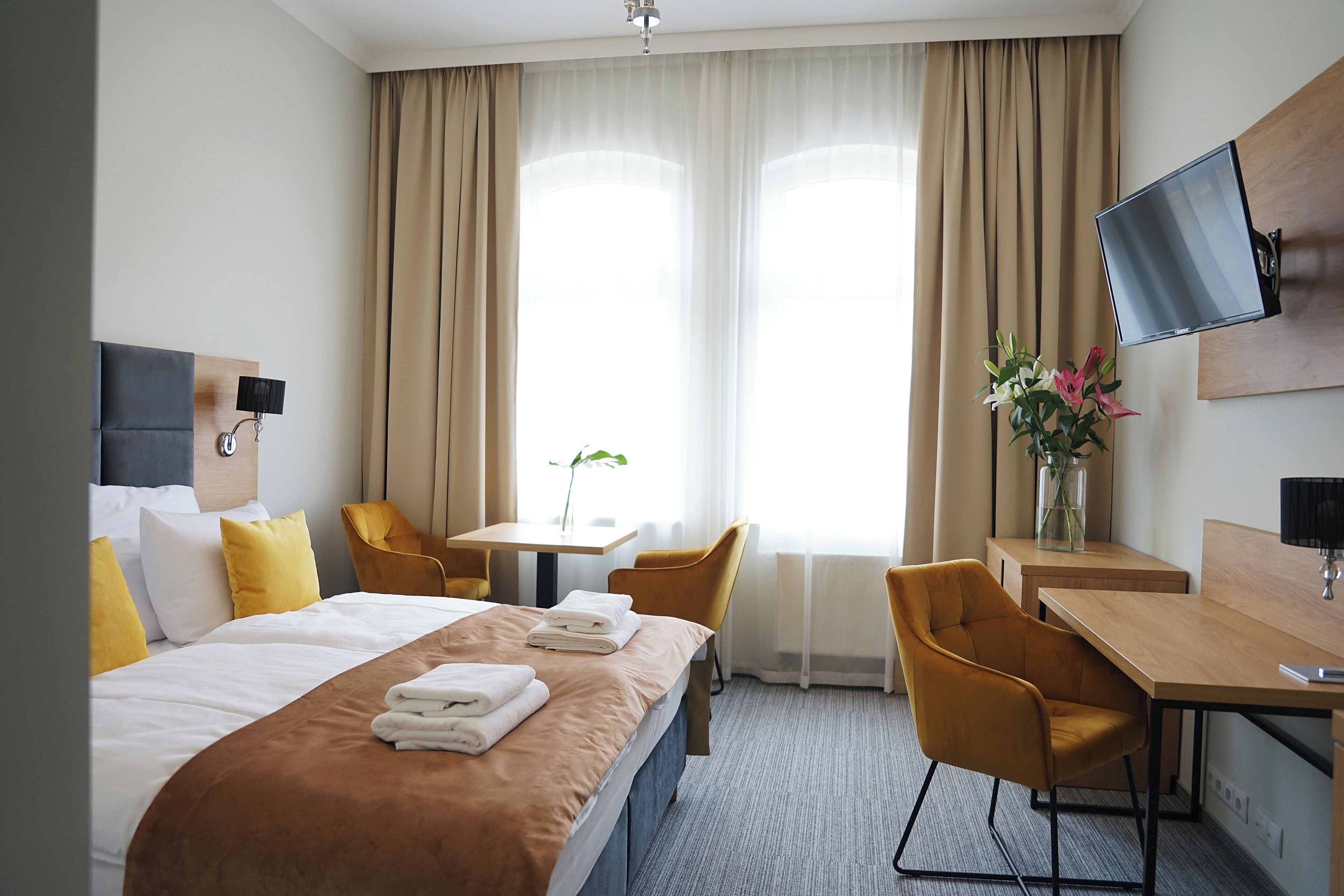 A hotel room with a bed, a desk, a chair, a TV mounted on the wall, and a window with curtains.