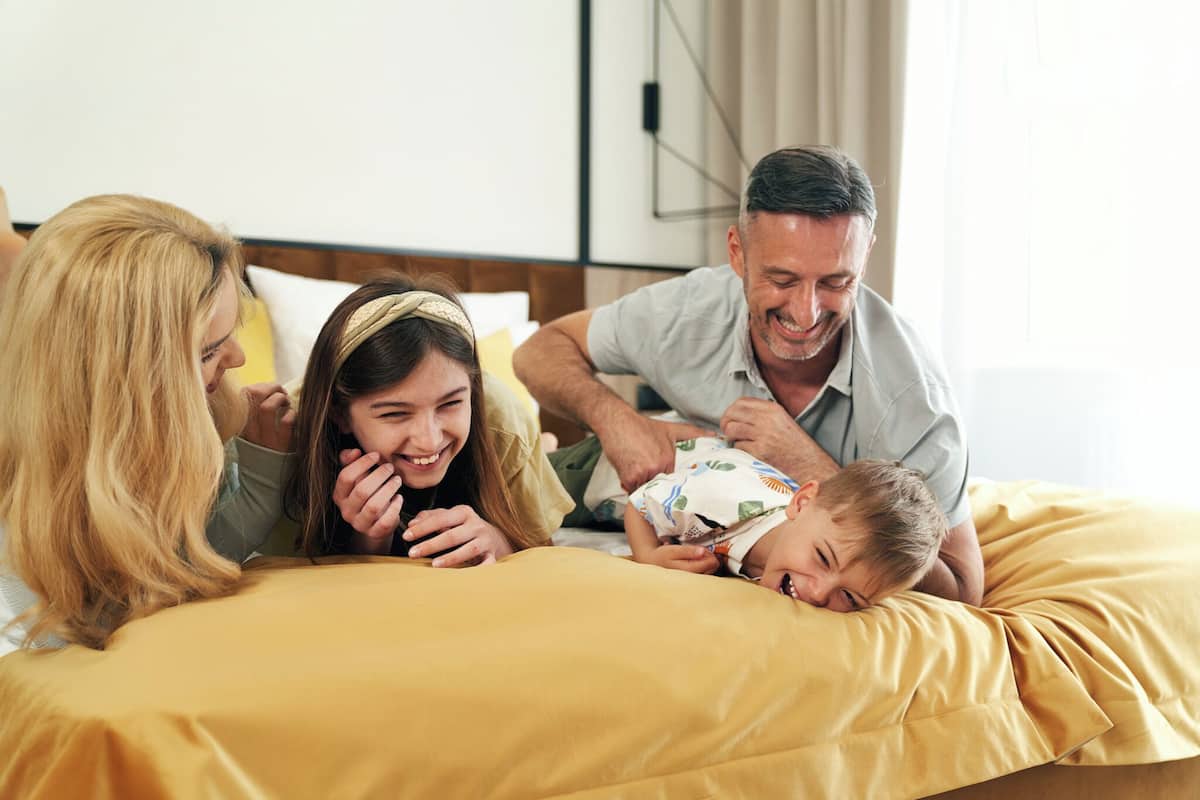 A family of four on a bed with a yellow blanket, a wooden headboard, and a window with curtains.