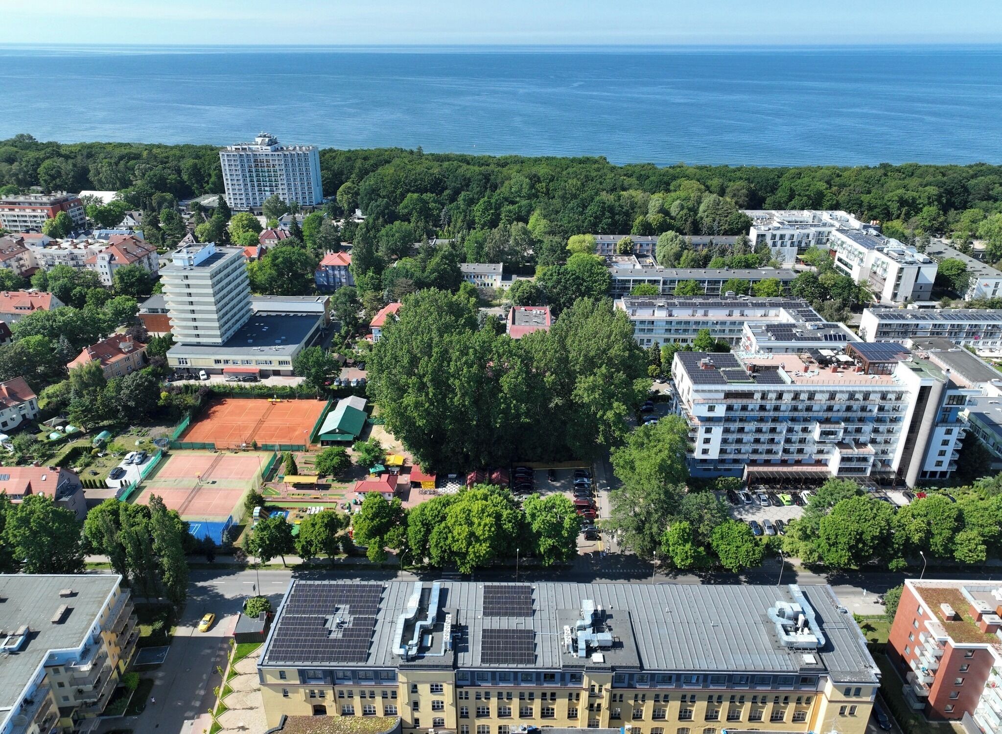 Vista aérea de una zona urbana costera con varios edificios, incluyendo un hotel grande con múltiples pisos y una terraza en la azotea.