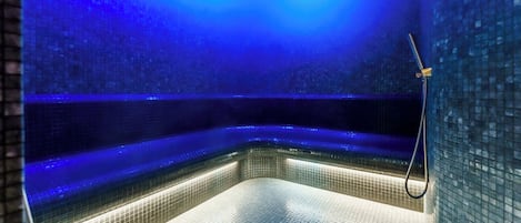 A modern, dimly lit indoor pool with a blue hue, featuring a corner shower area and tiled walls.
