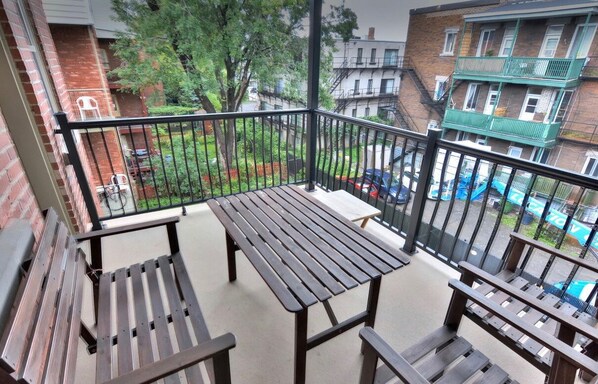 Un balcón con una mesa y bancos de madera, con vista a una zona residencial con edificios de ladrillo y un coche azul.