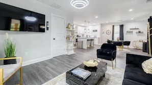 A modern living room with a black sofa, a coffee table, and a TV mounted on the wall. There is a kitchen area in the background with a dining table and chairs. - Atlanta's Hidden Gem: Modern 4BR/2BA House with Grill and Outdoor Space! (Atlanta)