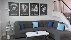 Ein modernes Wohnzimmer mit einer grauen Eckcouch, einem weißen Couchtisch und einem schwarzen Sitzsack. An der Wand sind vier Filmplakate angebracht und rechts führt eine Treppe nach oben.