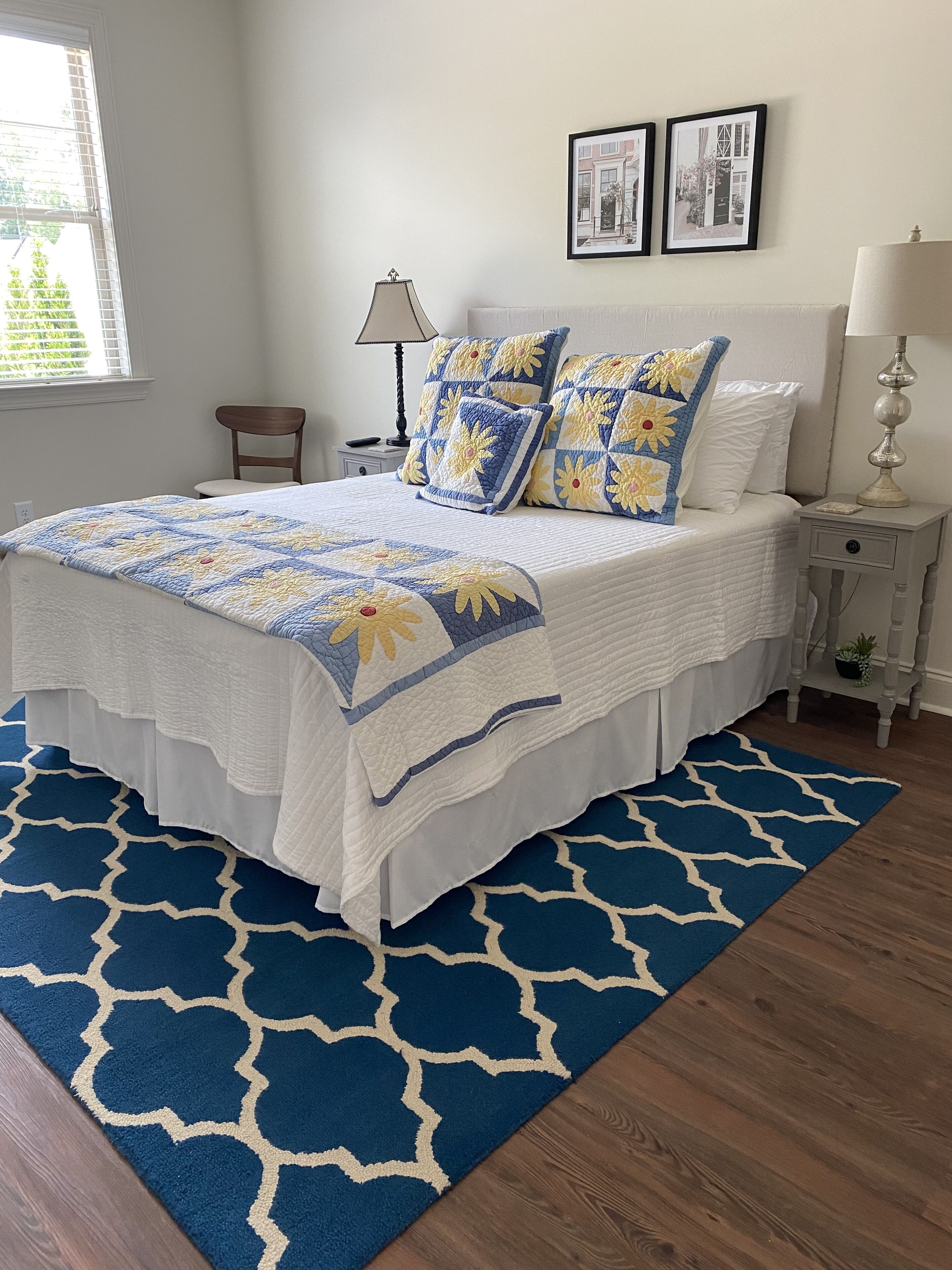 Una cama bien hecha con un edredón blanco y azul estampado y almohadas a juego, una mesita de noche blanca con una lámpara y una alfombra azul con diseño geométrico.