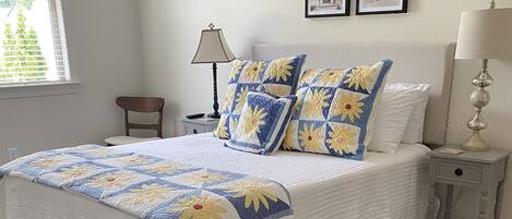 Una cama bien hecha con un edredón blanco y azul estampado y almohadas a juego, una mesita de noche blanca con una lámpara y una alfombra azul con diseño geométrico.