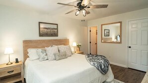 Une chambre à coucher avec un lit, des tables de chevet, un ventilateur de plafond, un tableau encadré et une porte.