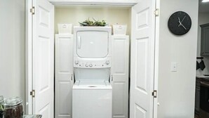 Une buanderie équipée d’un lave-linge et d’un sèche-linge à chargement frontal, d’une horloge au mur et d’une petite plante posée sur le dessus du sèche-linge.