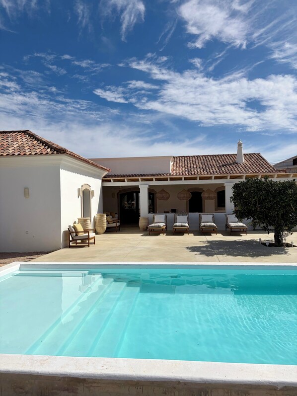 A poolside area with a clear blue pool, white walls, and a tiled roof. There are lounge chairs and a small tree.