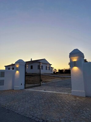A white building with a gated entrance, a cobblestone driveway, and two lighted pillars.