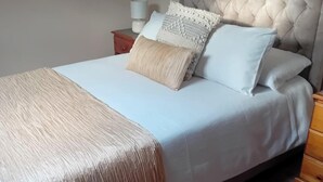 Un lit bien fait, avec une tête de lit grise, un plaid beige et plusieurs coussins.