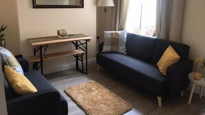 Un salon avec un canapé foncé, un fauteuil plus petit, une table en bois avec une lampe et un grand miroir au mur.