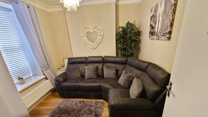 Un salon avec un canapé d’angle en cuir foncé, un petit tapis, un élément de décoration murale en forme de cœur et une plante en pot.