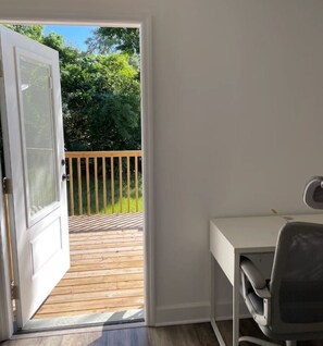A room with a white desk and chair, a sliding glass door leading to a wooden deck with a railing, and a view of greenery outside. - Brand new house Walking distance to Busch Gardens (Tampa)
