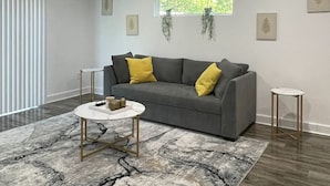Ein modernes Wohnzimmer mit einem grauen Sofa, gelben Kissen, einem Marmortisch und einem großen Fenster mit hängenden Pflanzen.