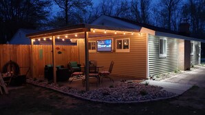Un área de estar al aire libre techada con una pantalla de televisión, piso de madera y luces colgantes.