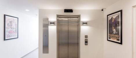 A modern elevator with a metallic finish, flanked by two wall-mounted light fixtures and a security camera above.