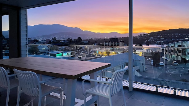 A balcony with a table and chairs, overlooking a cityscape with buildings and mountains during sunset. - Little Island Apartments (Bellerive)