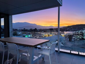 A balcony with a table and chairs, overlooking a cityscape with buildings and mountains during sunset. - Little Island Apartments (Bellerive)