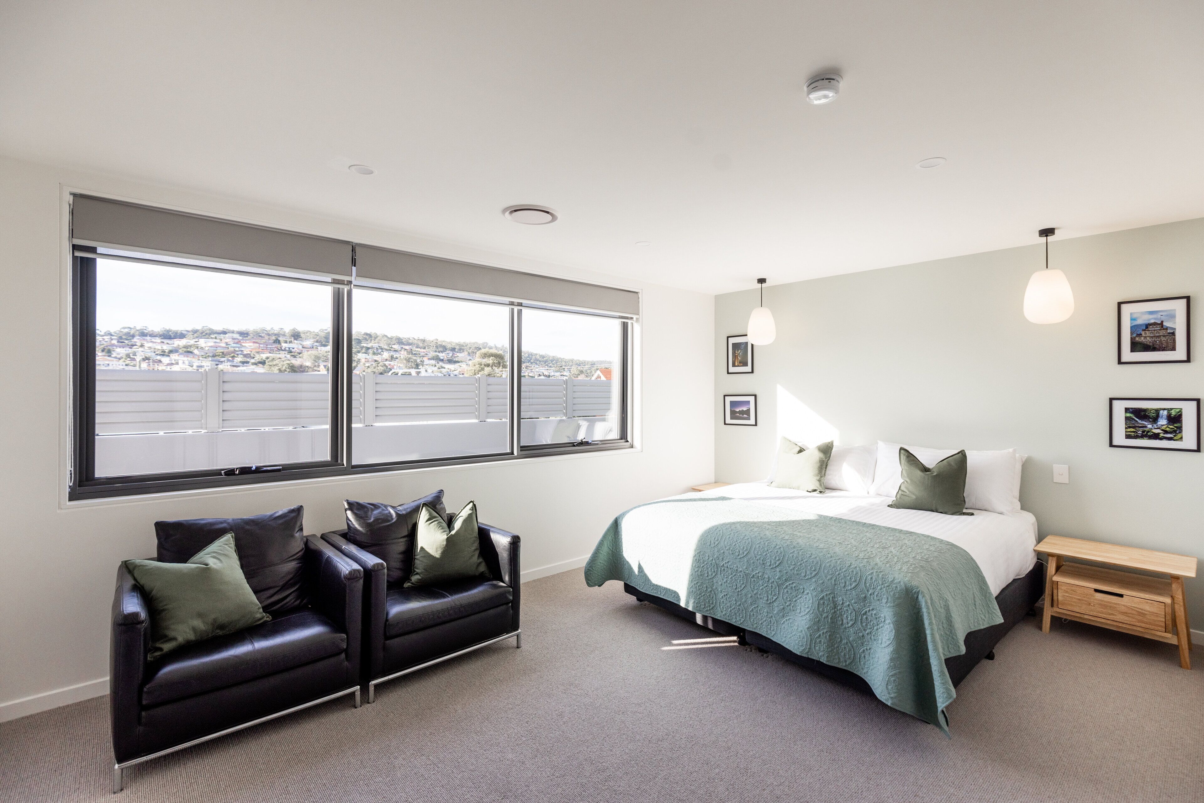 A modern bedroom with a large window, a bed with a green bedspread, a black leather sofa, and a wooden nightstand.
