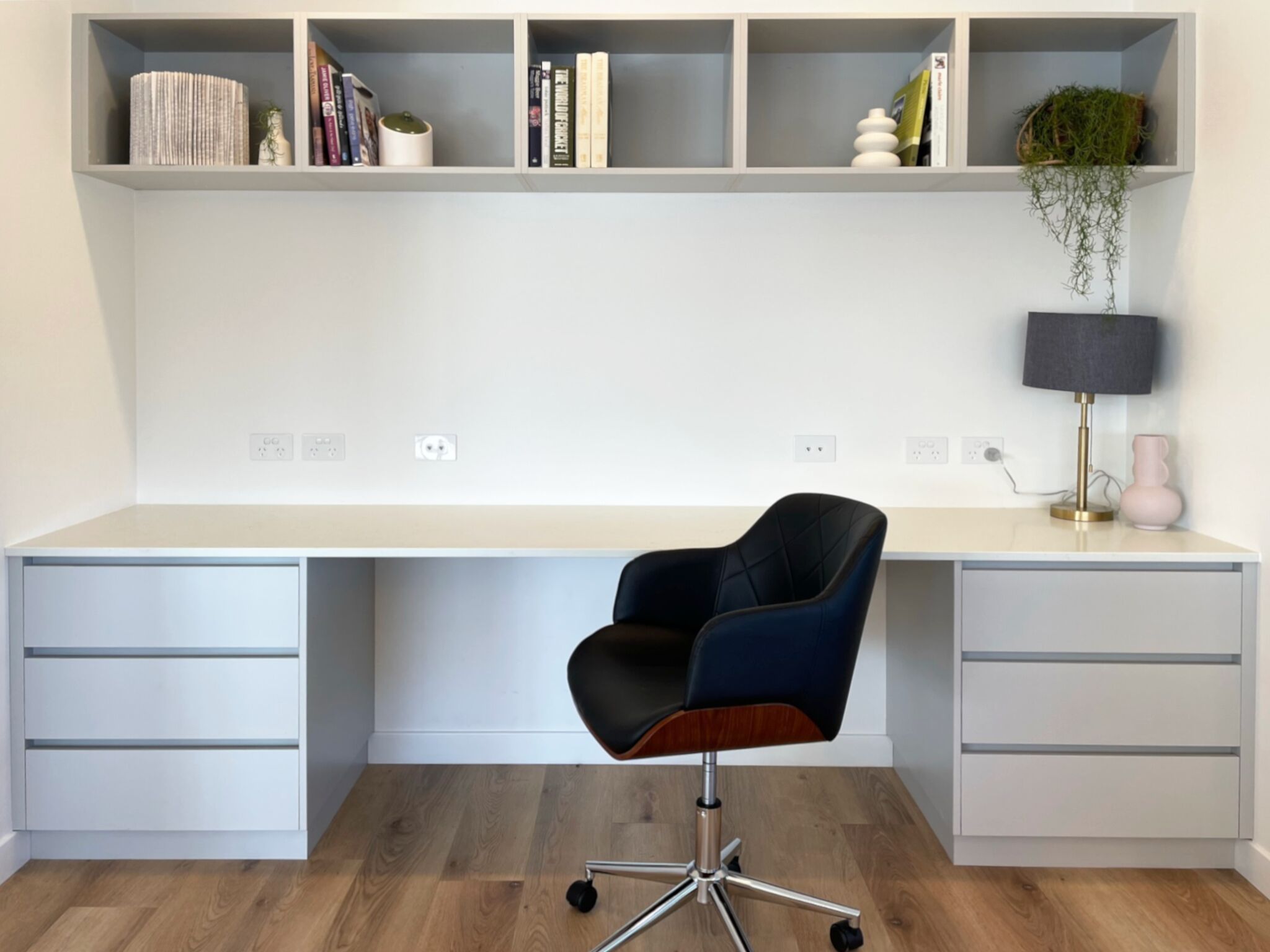 Un espacio de trabajo moderno con un escritorio grande, una silla de oficina negra y una estantería con libros y objetos decorativos.