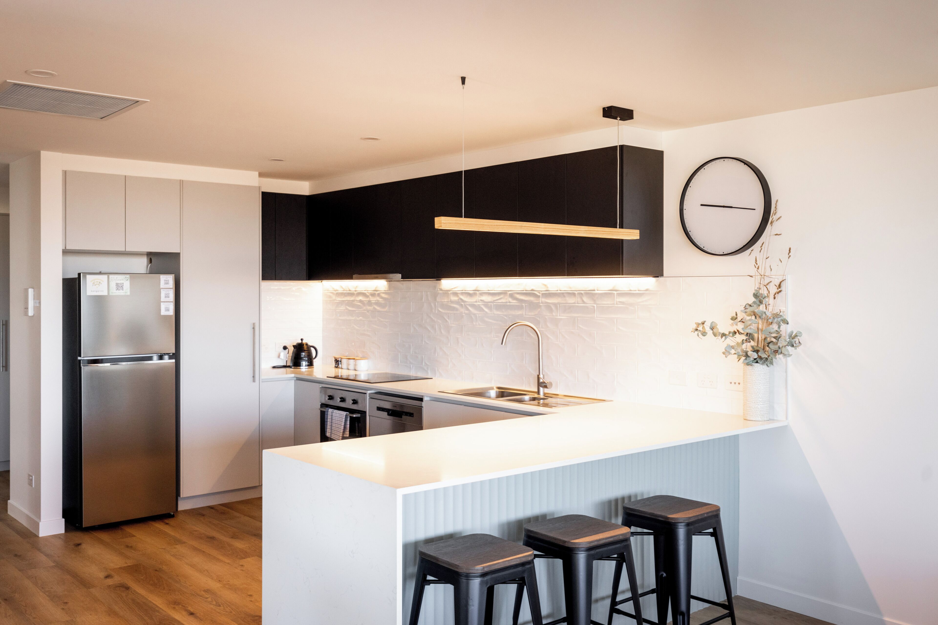 A modern kitchen with a stainless steel refrigerator, a built-in oven, and a sink. There are three black bar stools and a potted plant on the counter.