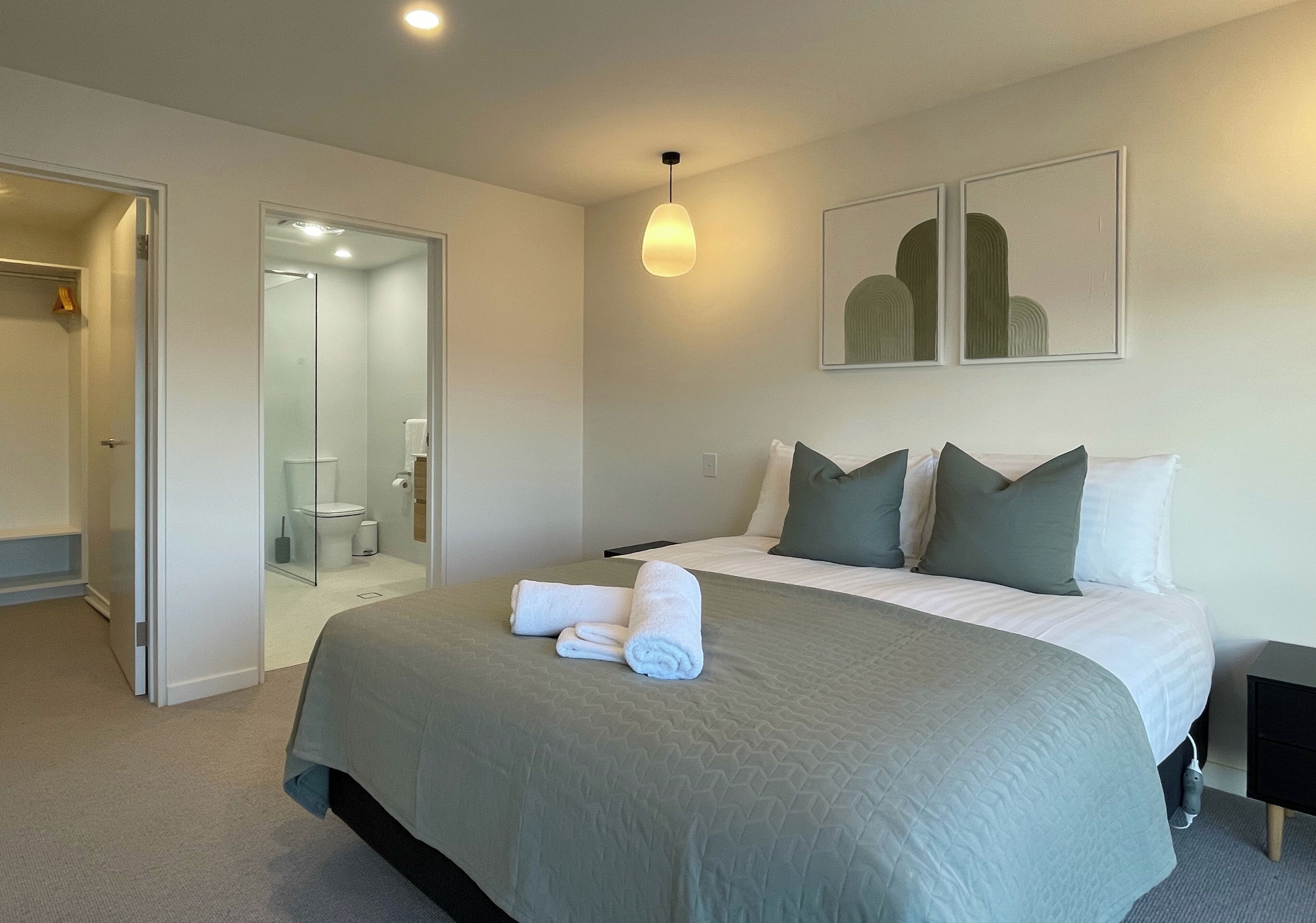 Habitación de hotel con una cama grande, una mesita de noche, una lámpara de pared y una puerta que da al baño.