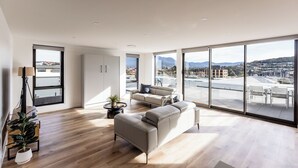 A spacious living area with a large window offering a city view, a sofa, a coffee table, and a potted plant. - Little Island Apartments (Bellerive)