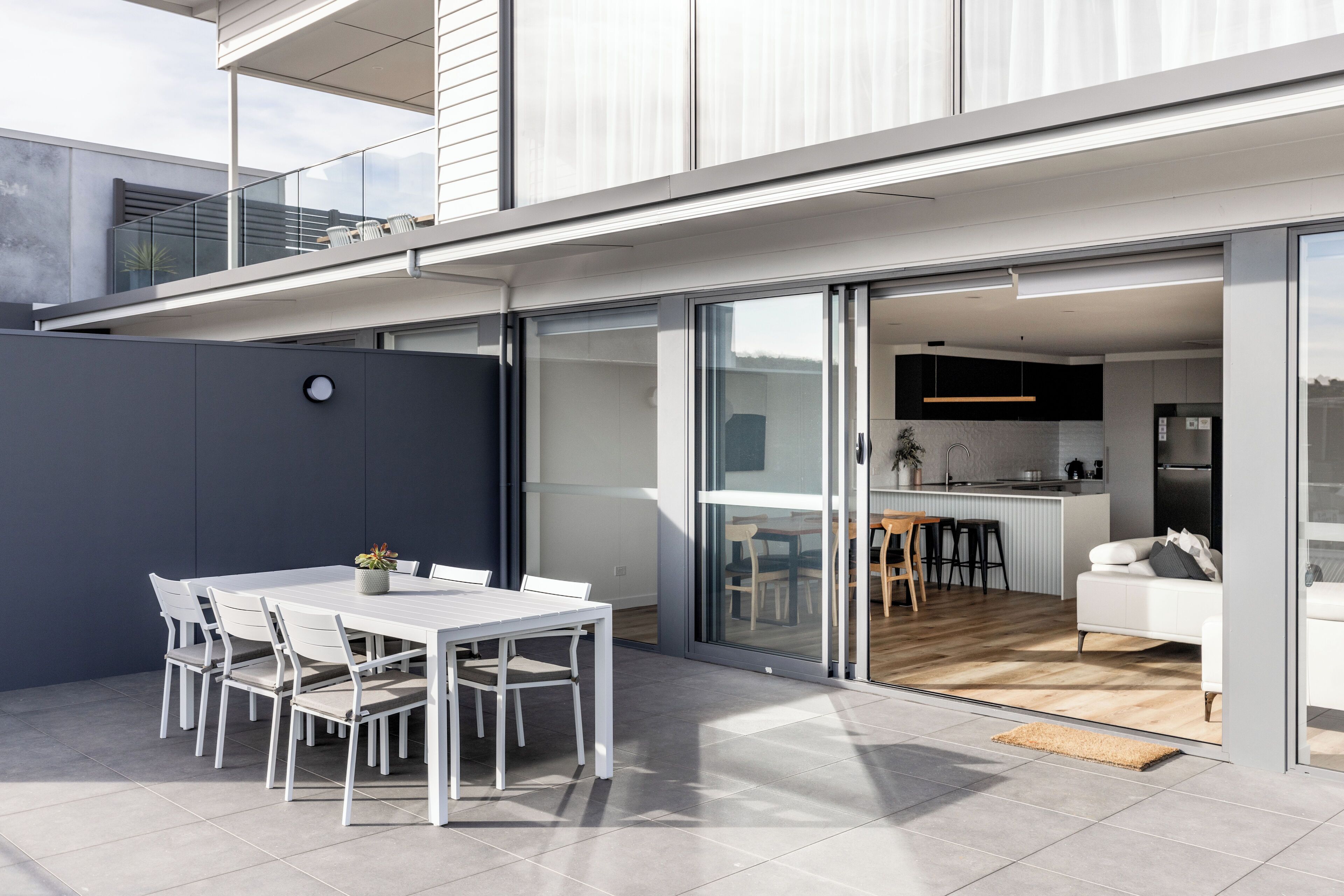 A modern residential building with a patio area featuring a dining table and chairs, a kitchen visible through glass doors, and a living area with a sofa and a coffee table.