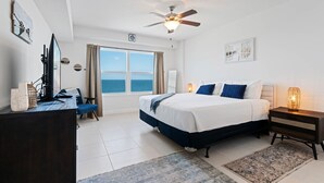Quarto com cama, mesas de cabeceira, escrivaninha, cadeira, ventilador de teto e vista para o oceano.
