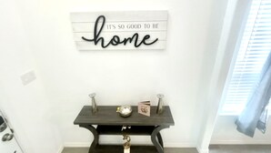 A modern interior with a dark wooden console table against a white wall, featuring a decorative sign above that reads "IT'S SO GOOD TO BE home." - DREAM LAND (Porter)