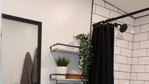 Un baño moderno con un lavabo blanco, un grifo negro y un inodoro blanco. Hay una cortina de ducha negra, una pequeña planta en maceta sobre una repisa y una alfombra de baño negra en el suelo.