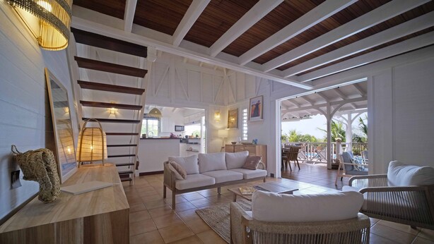A spacious living area with a sofa, chairs, and a coffee table. There is a kitchen in the background and a patio area with outdoor seating.