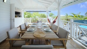 A covered outdoor dining area with a wooden table set for four, surrounded by cushioned chairs, and a view of a swimming pool and greenery in the background.