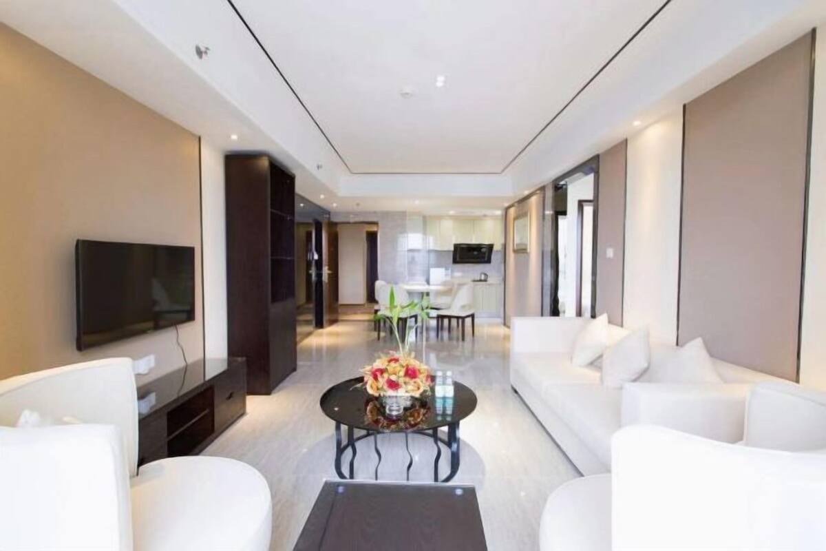 Een moderne woonkamer met een flatscreen televisie, witte banken, een salontafel met een bloemenarrangement en een keuken op de achtergrond.
