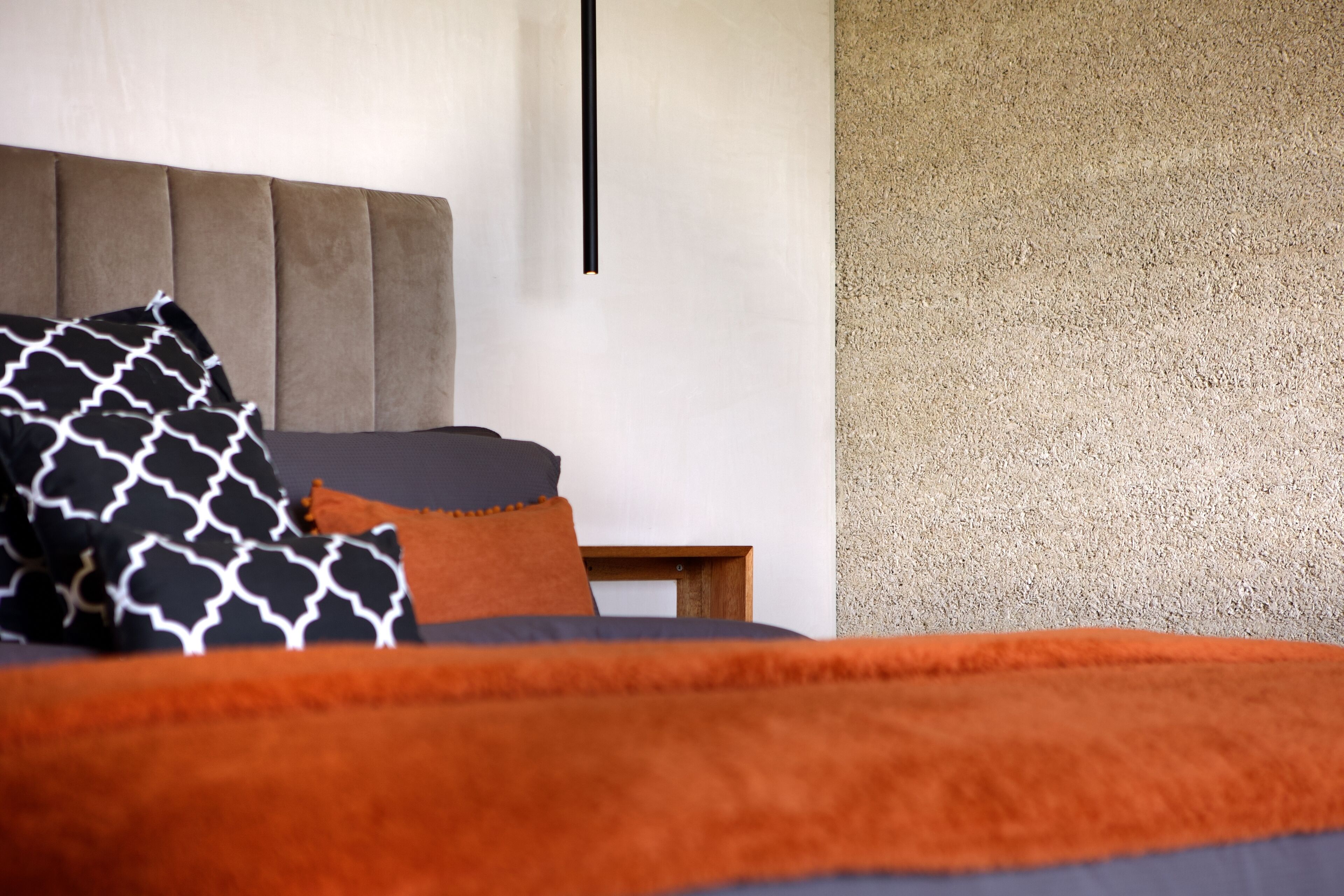 Een moderne slaapkamer met een gestoffeerd hoofdeinde, een bed met oranje en zwarte beddengoed, en een textuurwand aan de rechterkant.