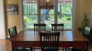 A dining area with a wooden table and chairs, large windows with a view of a garden, and a hanging light fixture.
