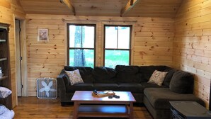 Ein gemütliches Blockhäuschen-Interieur mit Holzbalkendecke, Deckenventilator, einer Couch, einem Couchtisch und einem Kaminofen.