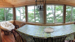 Eine Holzhütte mit einem runden Esstisch, bedeckt mit einer blumigen Tischdecke, umgeben von Stühlen und großen Fenstern mit Blick auf Bäume.