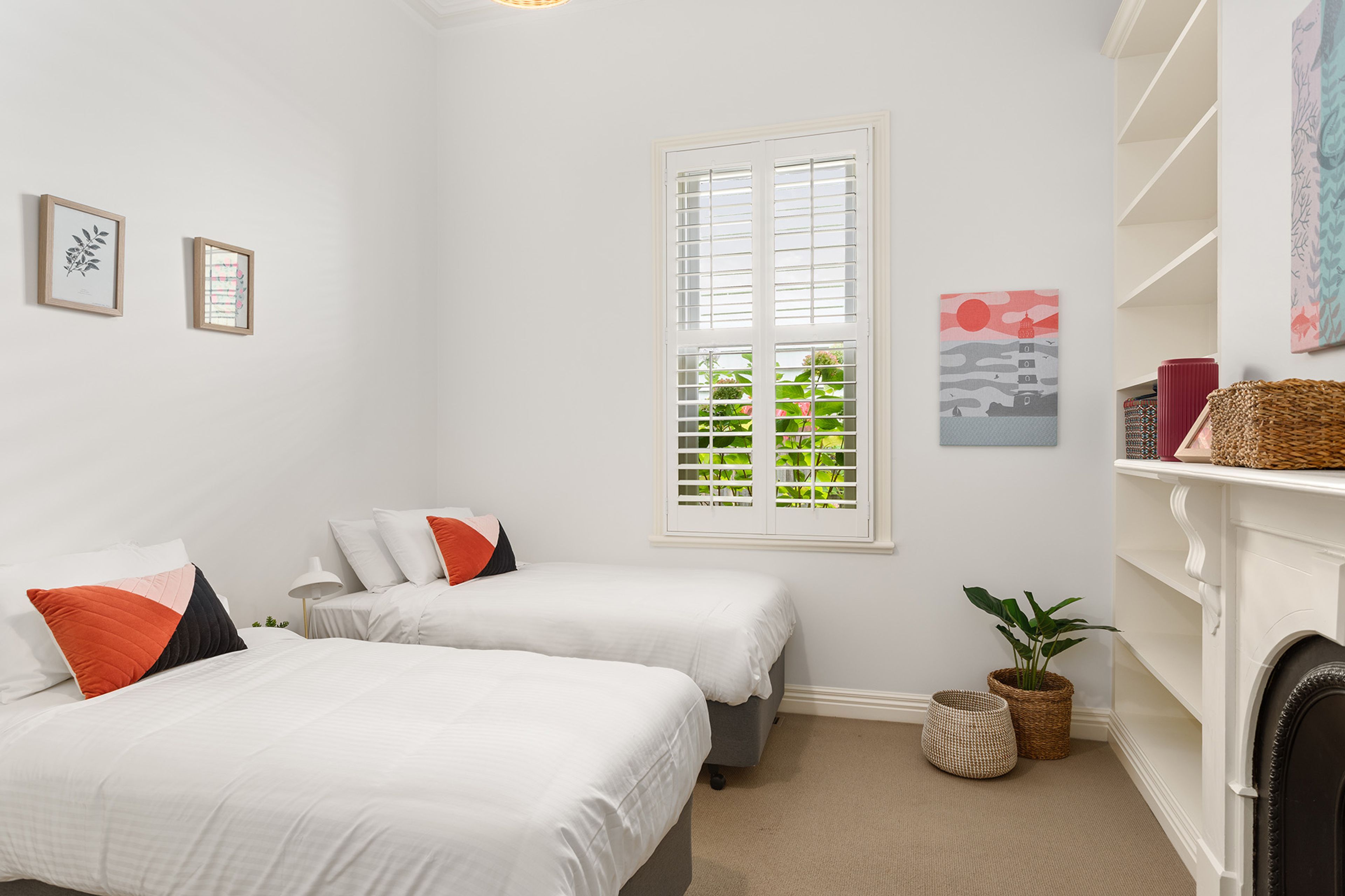 A bedroom with two beds, a window with blinds, a fireplace, and a shelf with decorative items.