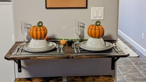 A dining table set for two with pumpkin decorations, a framed architectural drawing on the wall, and a modern kitchen in the background.