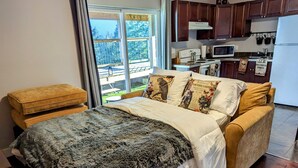 A bedroom with a large bed, a wooden bench, and a view of a kitchen and outdoor area.
