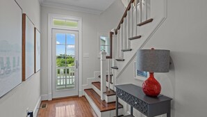 Un escalier avec des marches en bois et des rampes blanches, un vase rouge posé sur une console, et une porte donnant sur l’extérieur.