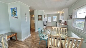 A spacious living area with a glass dining table, striped chairs, and a wooden bench. A kitchen is visible in the background with a window and a fire extinguisher. - The Reef Apartments Unit #2 - Downtown/5min walk to the beach! (Ocean City)