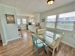 A dining area with a glass table and chairs, a kitchen in the background, and a view of the outside through large windows. - The Reef Apartments Unit #2 - Downtown/5min walk to the beach! (Ocean City)